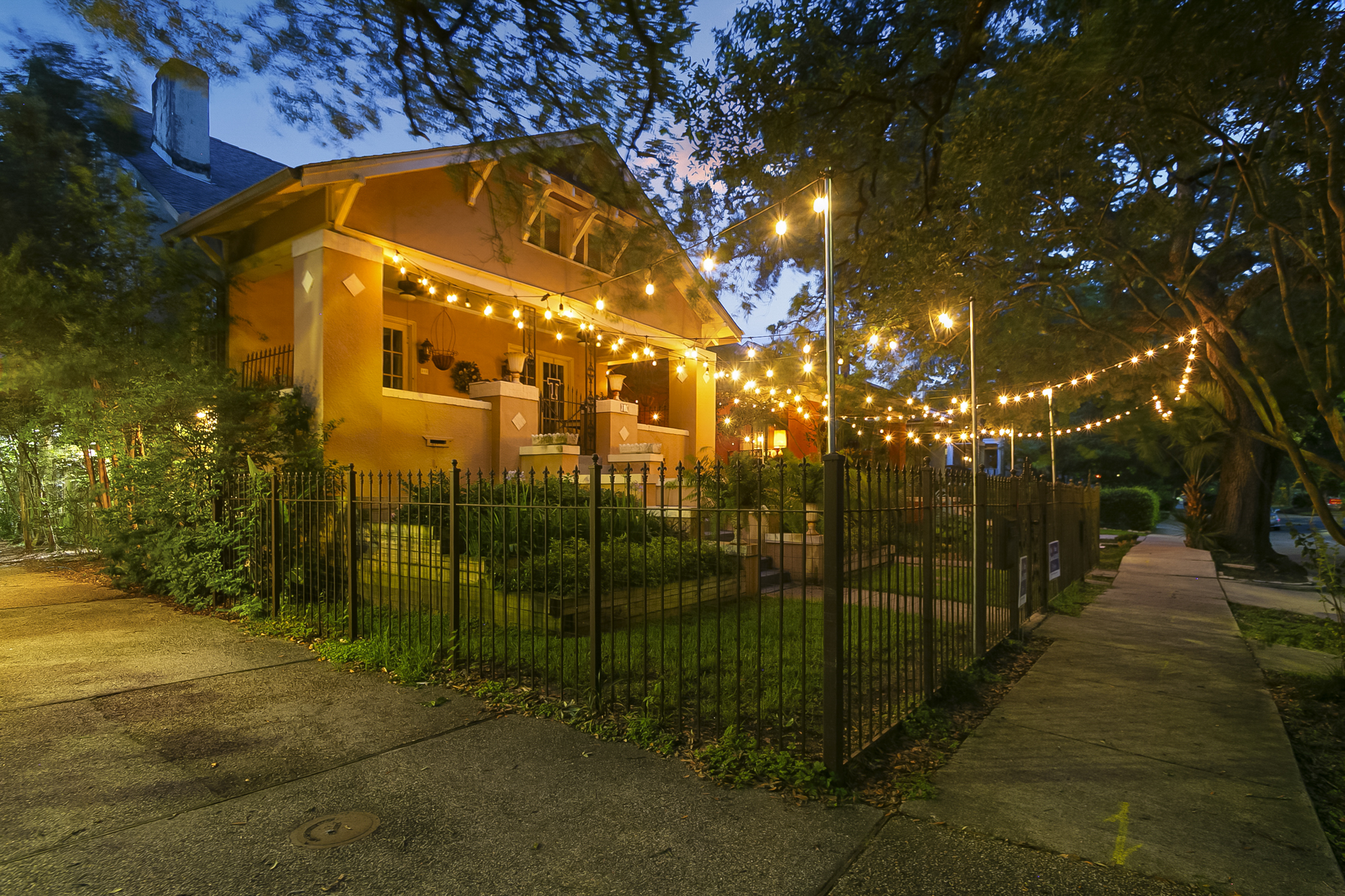 A New Orleans craftsman home glowing under string lights at dusk, framed by live oaks